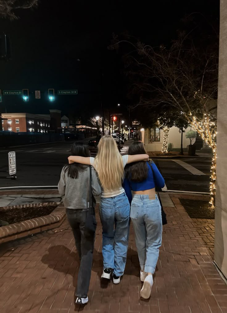 three girls walking down the sidewalk at night with their arms around each other's backs