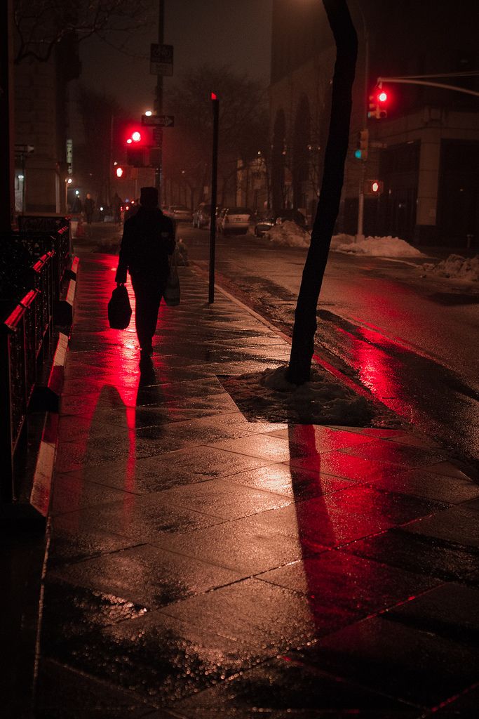 a person walking down the street in the rain at night with red traffic lights on