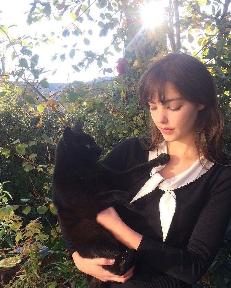 a woman holding a black cat in her arms while standing next to bushes and trees