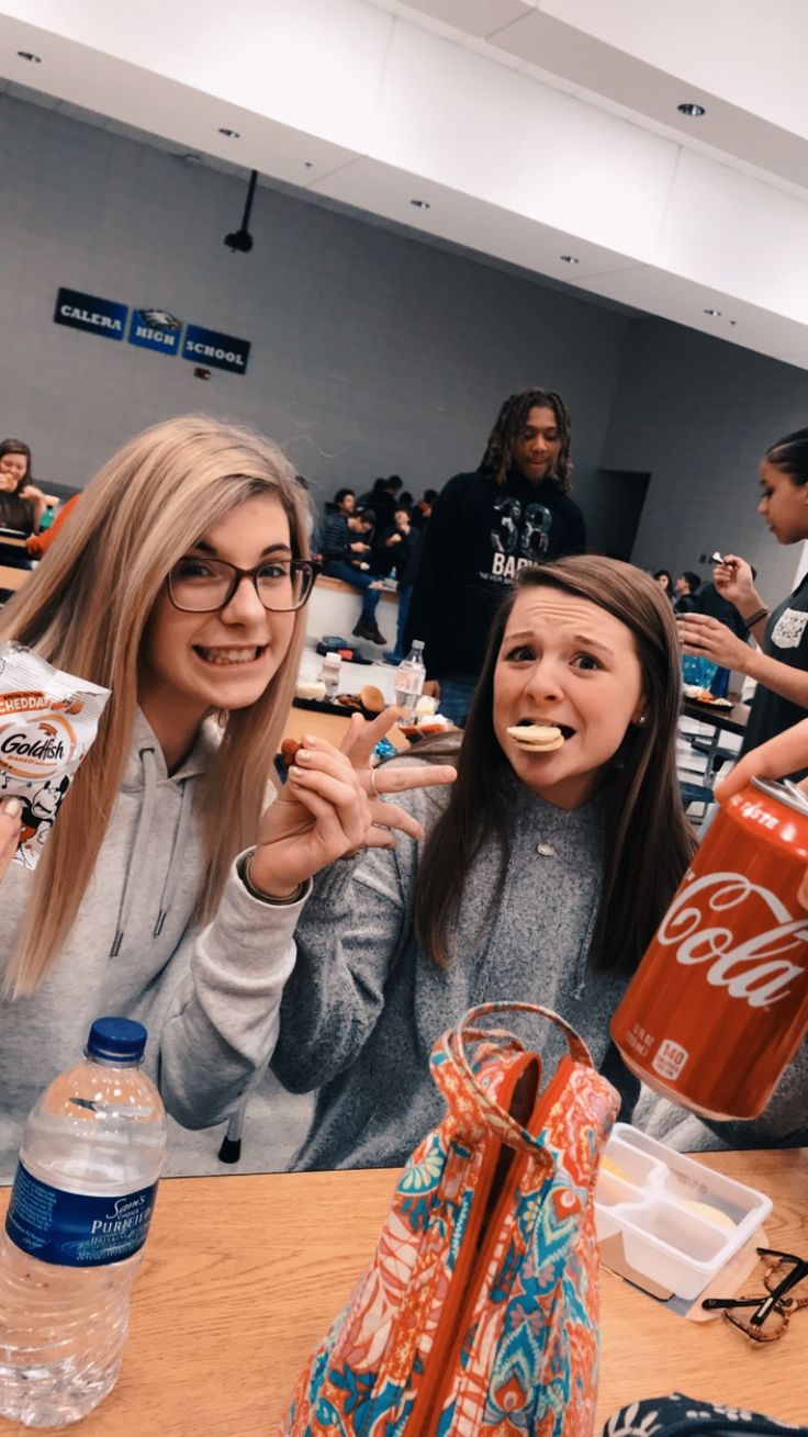 two women sitting at a table eating food and drinking sodas with other people in the background