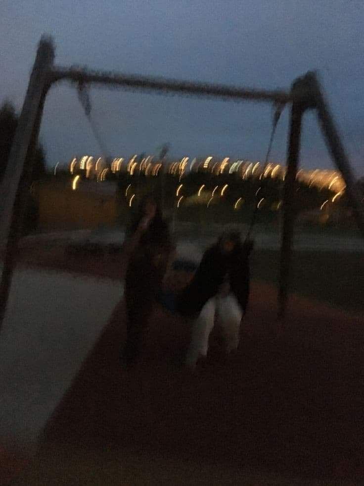 two people are playing on a swing set at night, with lights in the background