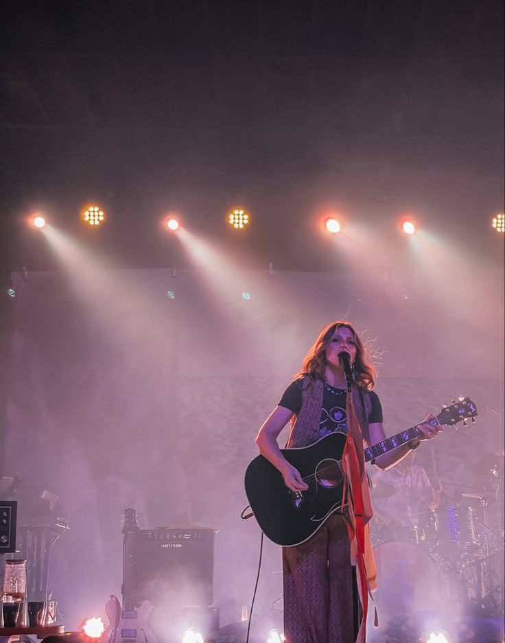 a woman standing on top of a stage with a guitar in front of her and lights behind her