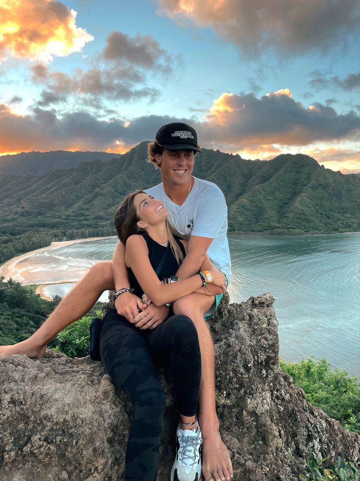 a man and woman sitting on top of a rock next to the ocean at sunset