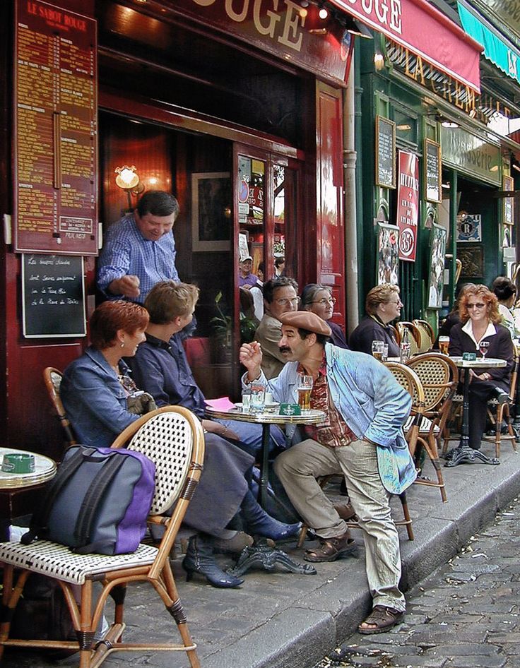 people are sitting at tables outside in front of a restaurant on the side of the street