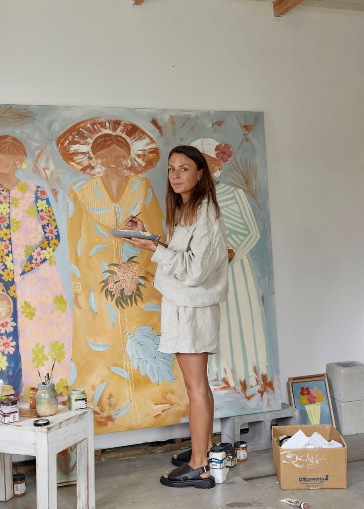 a woman is standing in front of a painting and holding a paintbrush while looking at it