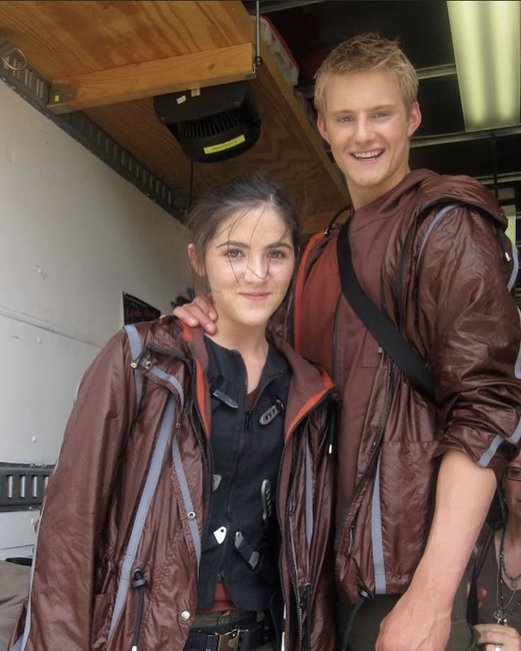 two people standing next to each other in front of a building and one is wearing a brown jacket