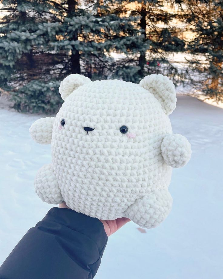 a hand holding up a crocheted stuffed animal in the snow with pine trees in the background