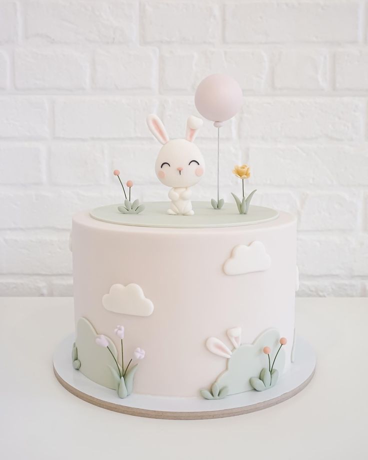 a white cake decorated with flowers and a bunny on top is sitting in front of a brick wall