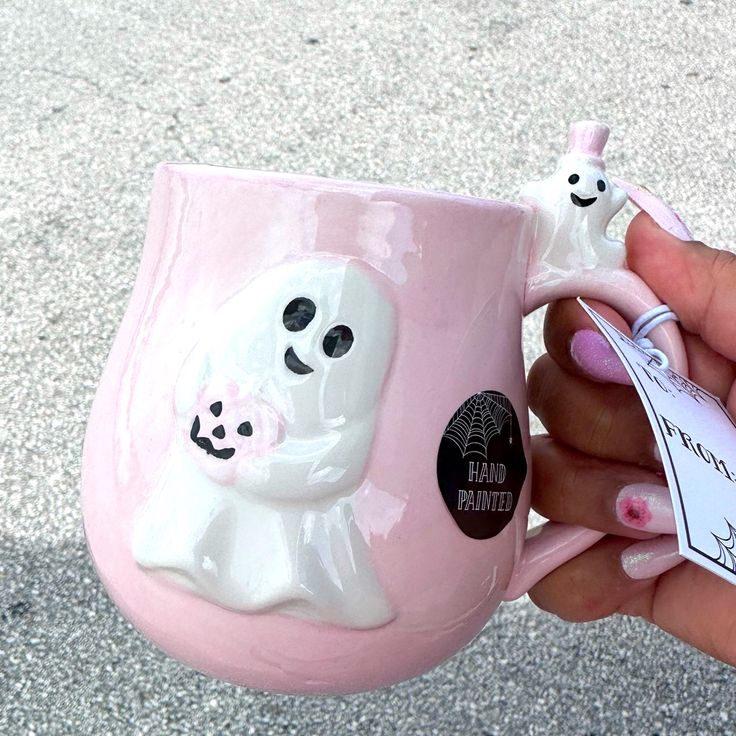 a hand holding a pink coffee mug with two ghost faces on it and a tag attached to the handle