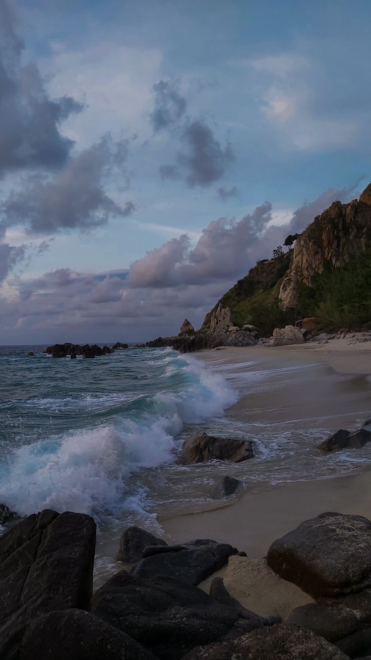 the ocean waves are crashing onto the rocks on the beach at dusk with an island in the background