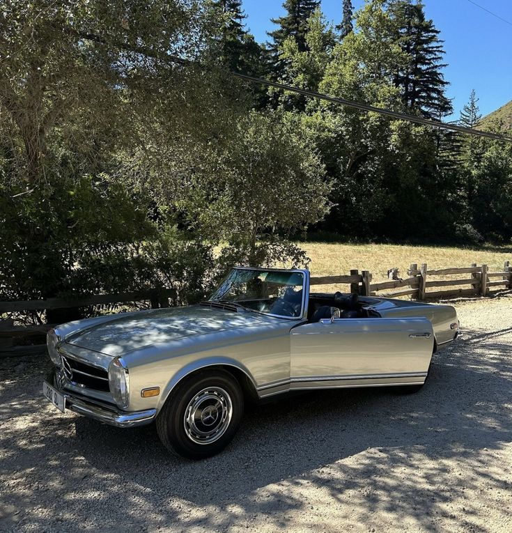 an old car is parked in the gravel near some trees and fenced in area