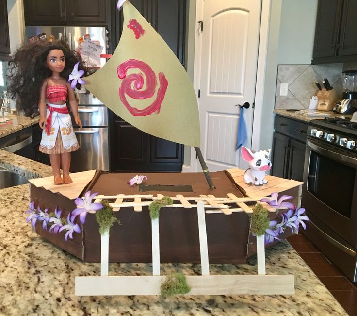 a doll is standing next to a cake in the shape of a sailboat on a kitchen counter