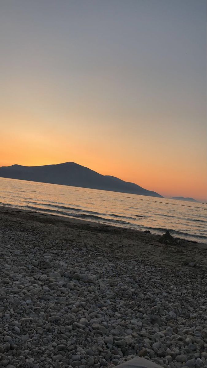 the sun is setting at the beach with rocks in front of it and mountains in the distance