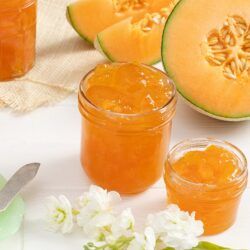 two jars filled with jam next to sliced melon and flowers on a white surface