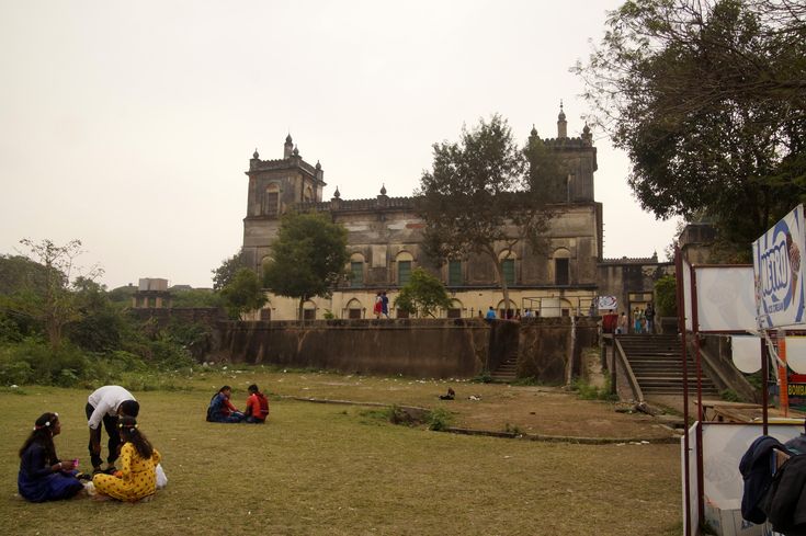 two women sitting on the grass in front of an old building with people standing around