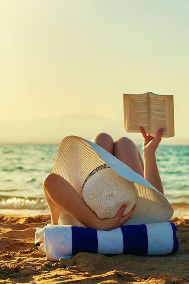 a woman laying on top of a beach next to the ocean holding an open book