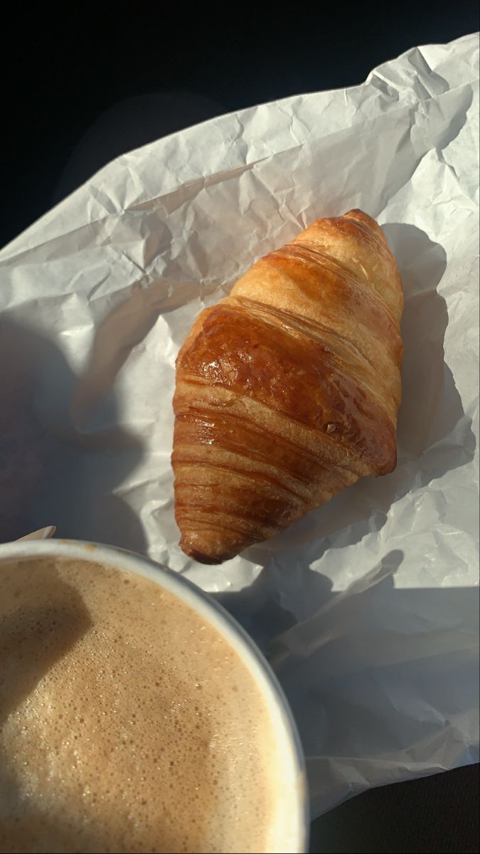 a croissant sitting on top of a piece of paper next to a cup of coffee