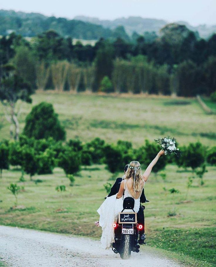 a bride and groom riding on a motorcycle down a dirt road with their arms in the air