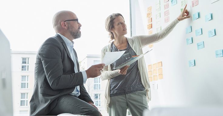 two people standing in front of a whiteboard with sticky notes on it and one person pointing at the board