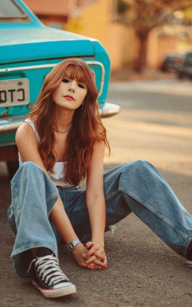 a woman sitting on the ground next to a blue car with her hands in her pockets