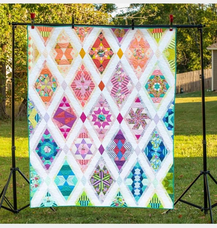 a colorful quilt hanging on a black stand in the grass with trees and houses behind it