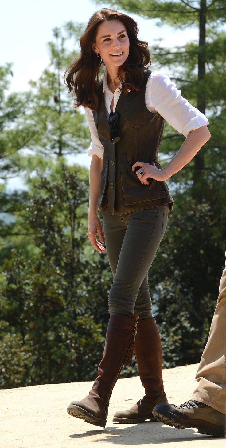 a woman standing on top of a hill wearing boots and a vest with her hands on her hips