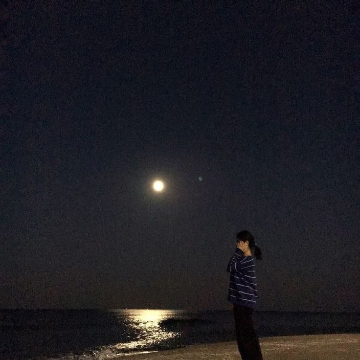 a woman is standing on the beach at night looking up at the moon and stars