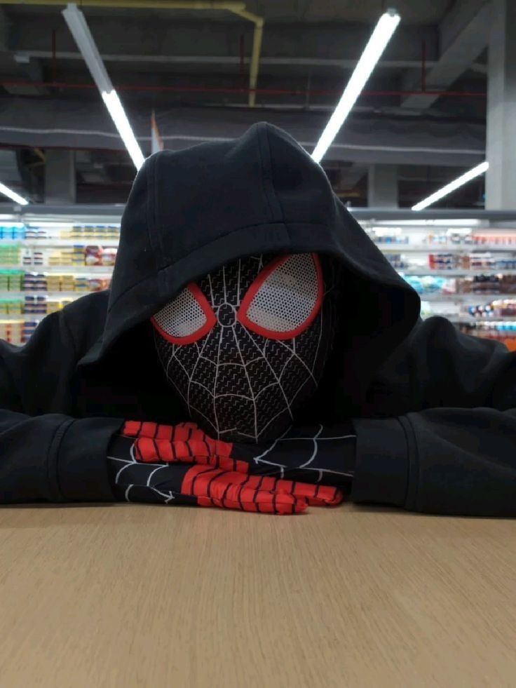 a person wearing a spider man mask sitting at a table in a grocery store with their hands on his head