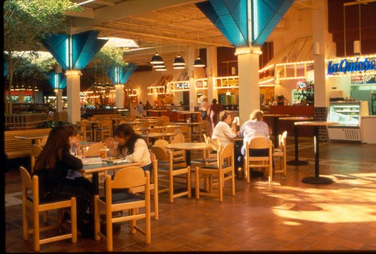 people sitting at tables in a restaurant with blue lights on the ceiling and windows above them