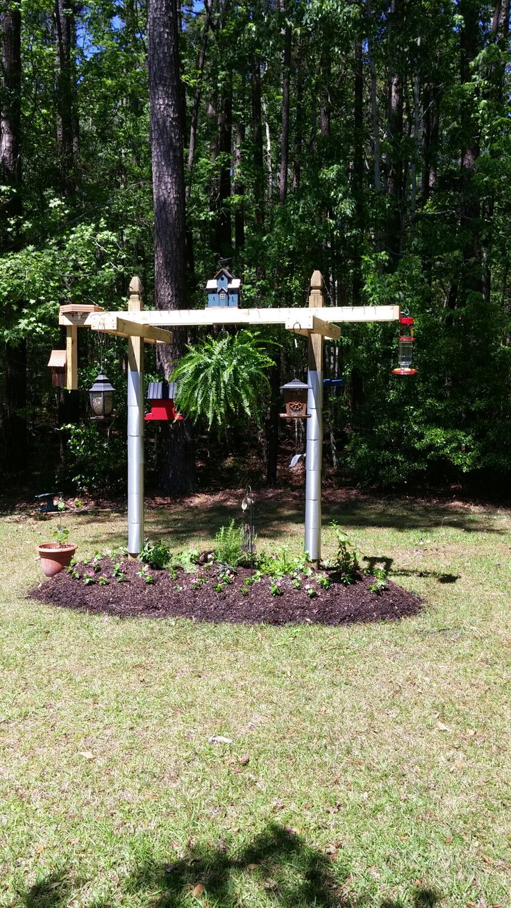 a bird feeder in the middle of a flower bed with trees and bushes behind it