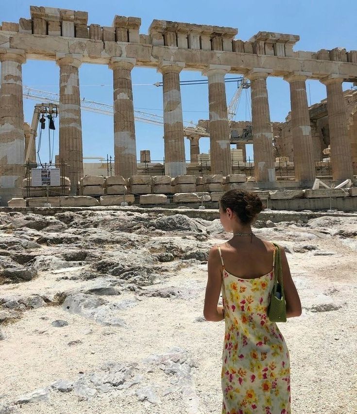 a woman standing in front of the ruins of an ancient city, looking up at the sky