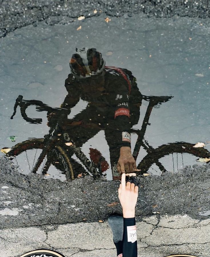 the reflection of a man riding a bike in a puddle with his hand on the ground