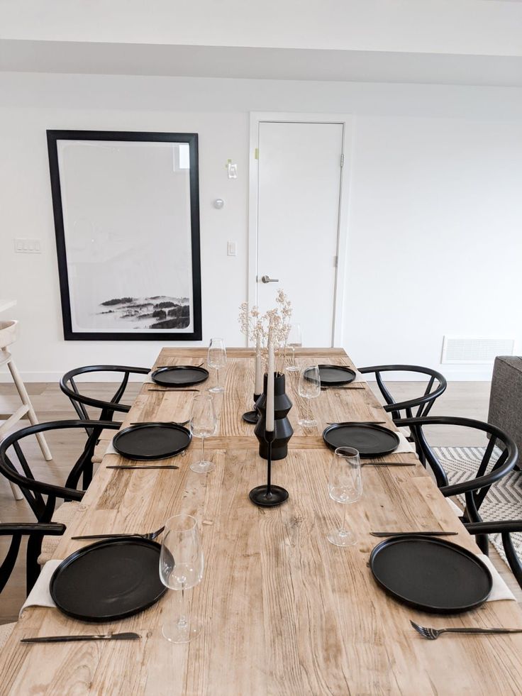 a wooden table with black plates and silverware on it in a white walled dining room