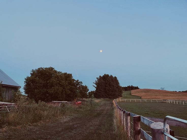 an old barn sits on the side of a dirt road in front of a fence