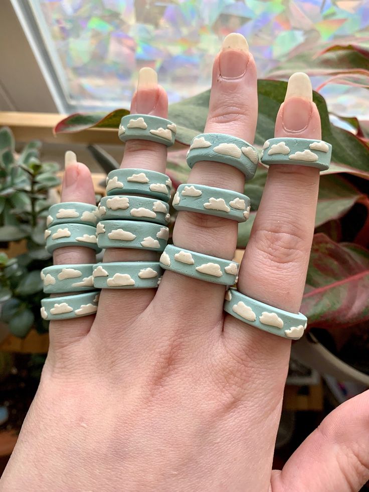 a woman's hand holding five rings with clouds painted on them, in front of a potted plant