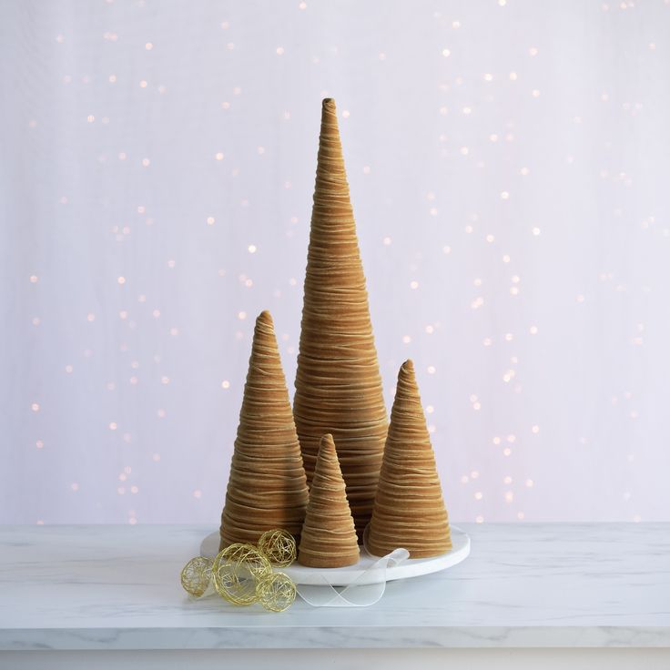 three small wooden christmas trees on a white plate with gold ribbon and lights in the background