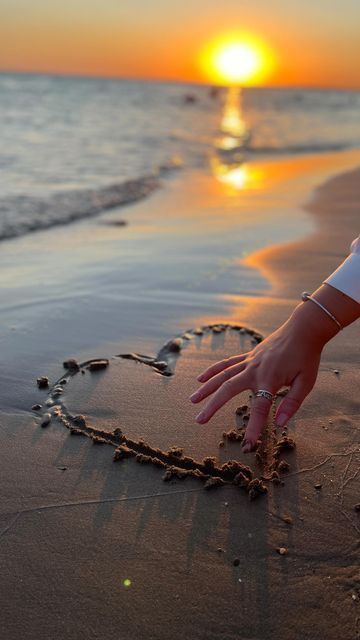 a person's hand on the beach making a heart shape with their fingers at sunset