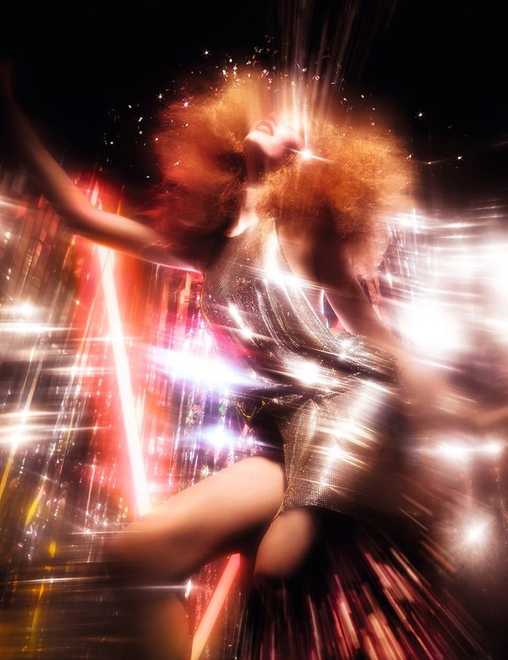 a woman with red hair is dancing in front of bright lights and sparkles on her body