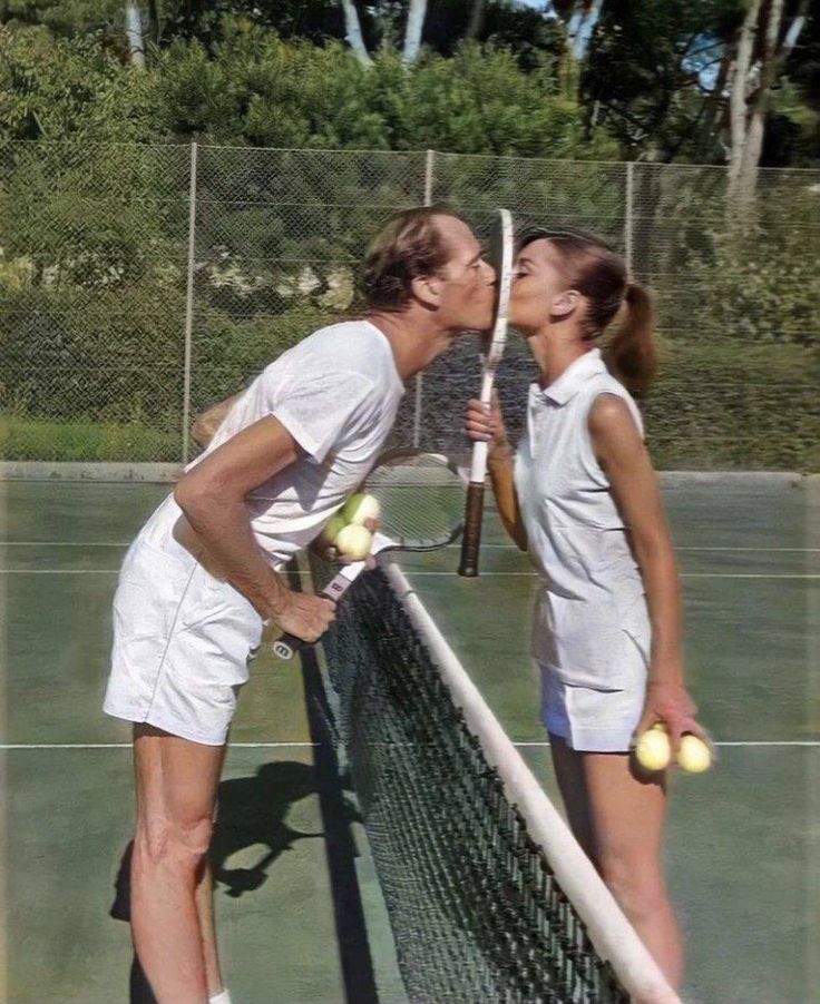 two people standing on a tennis court with rackets and balls in their hands, kissing