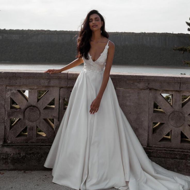 a woman in a white wedding dress standing on a stone wall near the water and trees