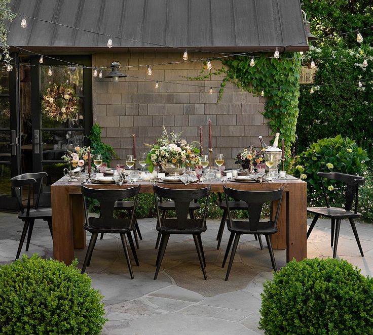 an outdoor dining table set for six with chairs around it and greenery in the background