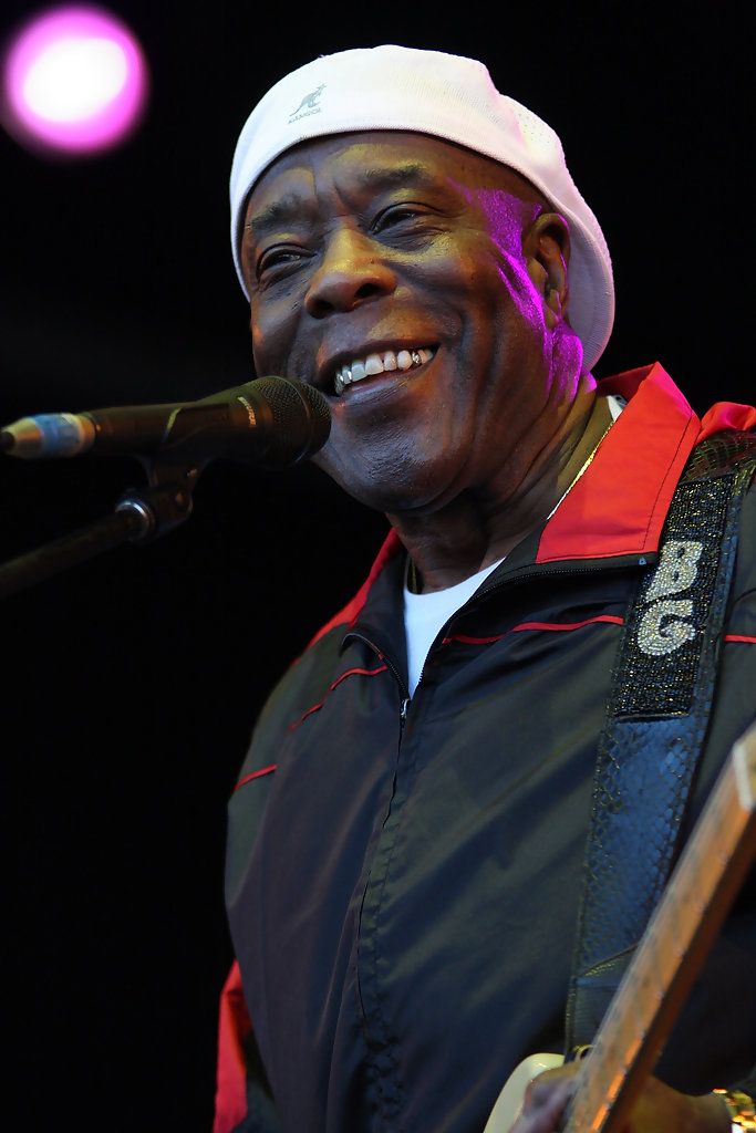 a man with a white hat on singing into a microphone and holding a guitar in his right hand