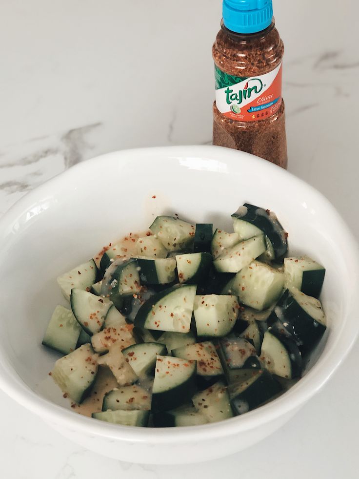 a white bowl filled with sliced cucumbers next to a bottle of seasoning