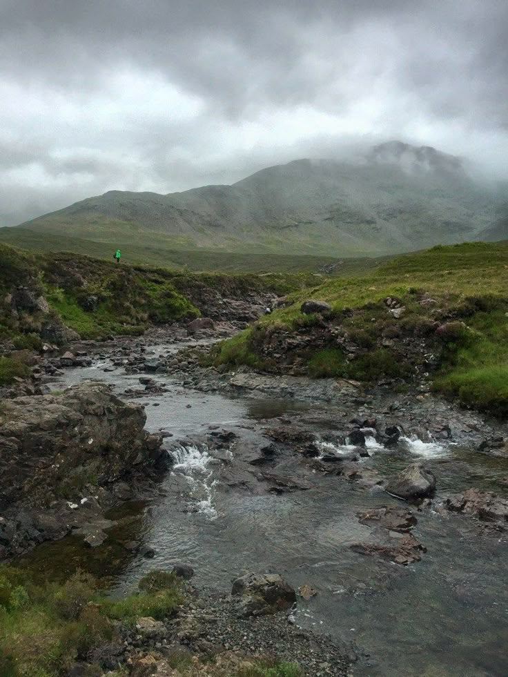 a small stream running through a lush green hillside covered in foggy clouds and grass