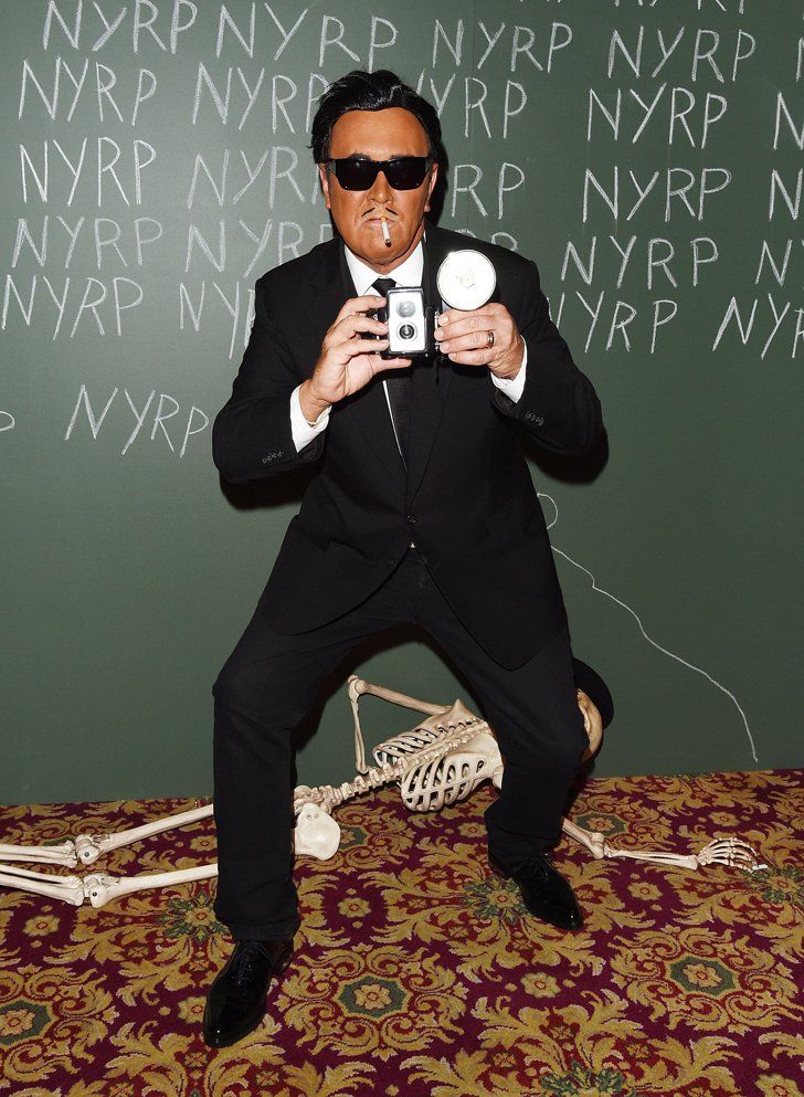 a man in a suit and sunglasses is holding a camera while sitting on a skeleton