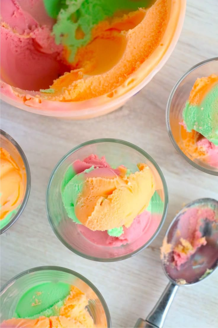 four bowls filled with colorful ice cream on top of a white table next to spoons