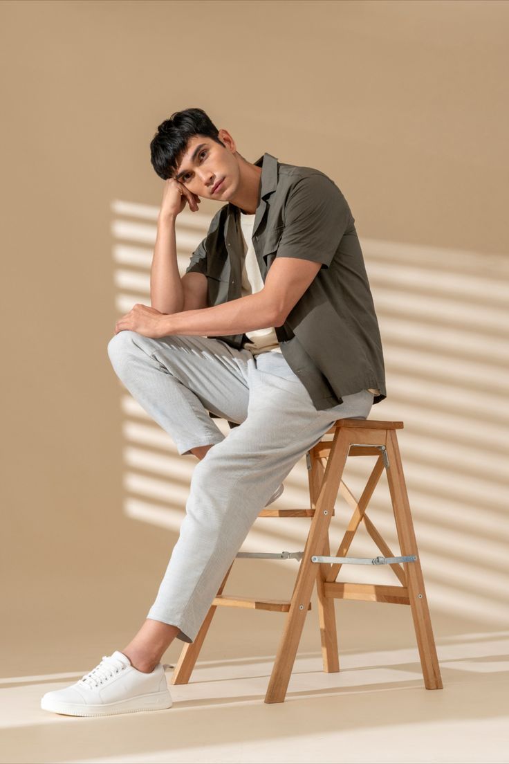 a young man sitting on top of a wooden stool next to a white wall with his hand under his chin