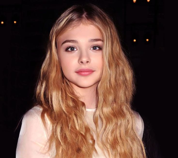 a girl with long blonde hair and blue eyes looks at the camera while wearing a white shirt