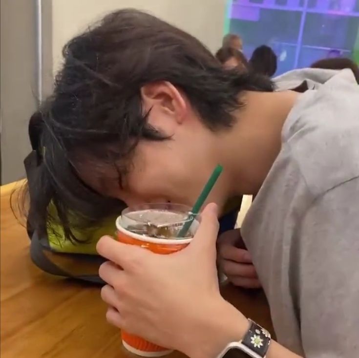 a young man drinking from a cup while leaning his head down on a table with other people in the background