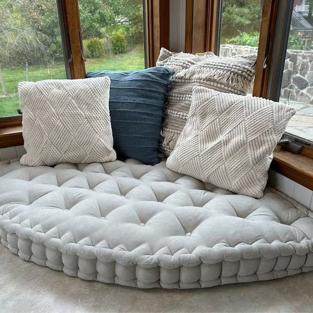 a round couch with four pillows on it in front of a window and some windows